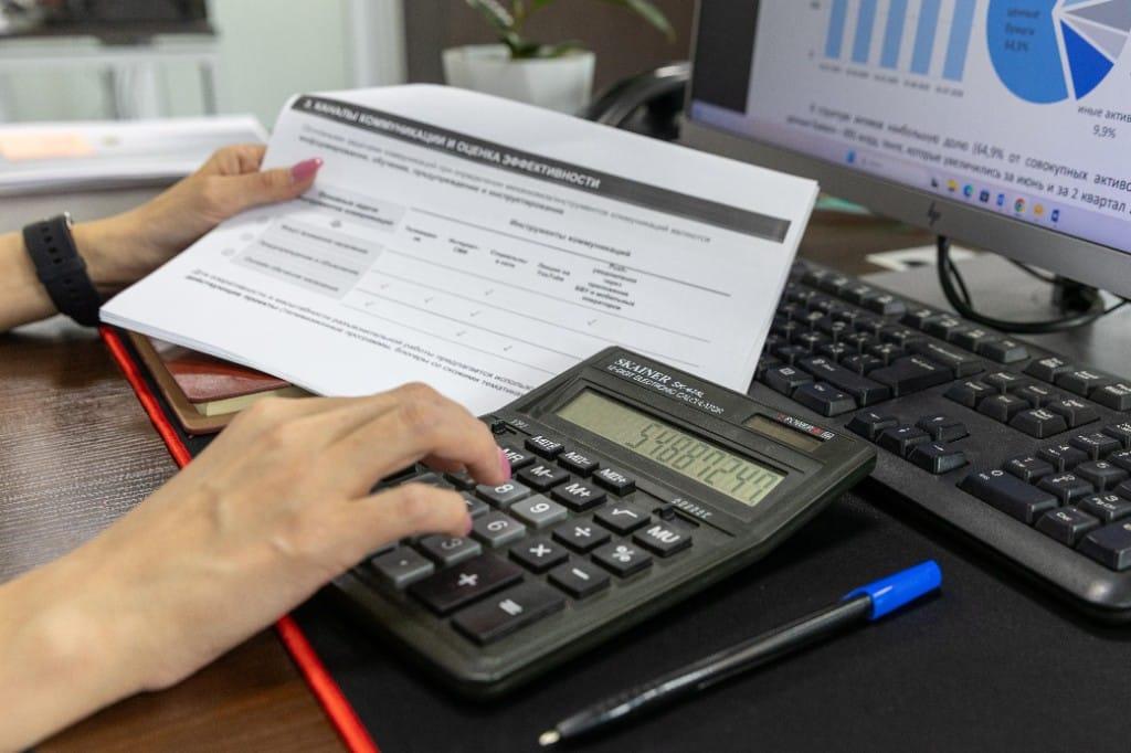 Financial analyst reviewing statements and using a calculator beside charts on a computer screen